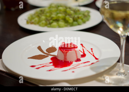 Elevato angolo di visione di un individuo che serve dei deliziosi dessert mousse guarnita con ricco rosso sciroppo fruttato su un ristorante tavolo con un bicchiere di vino bianco. Foto Stock