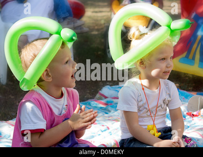 Due giovani bambini bionda indossare cappelli realizzato da ritorto palloncini verde seduti su un tappeto sul pavimento godendo di una festa di compleanno Foto Stock