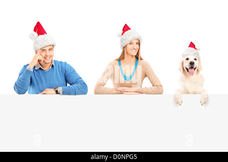 Coppia sorridente e cane con Babbo Natale cappelli in posa dietro un pannello vuoto Foto Stock