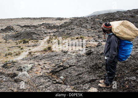 MONTE KILIMANJARO, Tanzania - Un portiere si ferma per un momento per riposare sul sentiero tra il Moir Hut Camp (13.660 piedi) e la Lava Tower (15.215 piedi) sul percorso Lemosho del monte Kilimangiaro. A questa altitudine, la zona brughiera lascia il posto al deserto alpino roccioso. Foto Stock