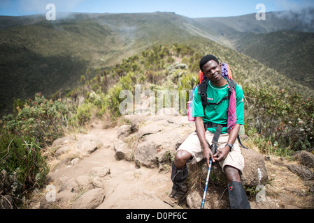 Guida del Monte Kilimangiaro Sentiero Lemosho Tanzania | MONTE KILIMANJARO, Tanzania — Una guida si riposa lungo il Sentiero Lemosho a circa 10.000 piedi (3.050 m) di altitudine tra Big Tree Camp e l’Altopiano di Shira. La vegetazione bassa visibile lungo la cresta caratterizza la zona di brughiera della montagna, una fascia ecologica che sostituisce le foreste montane inferiori. Questo ambiente semi-alpino si estende fino a 13.000 piedi (4.200 m), agendo come una transizione di acclimatamento prima che gli scalatori entrino nel deserto alpino sterile al di sotto della vetta a 19.341 piedi (5.895 m). Foto Stock