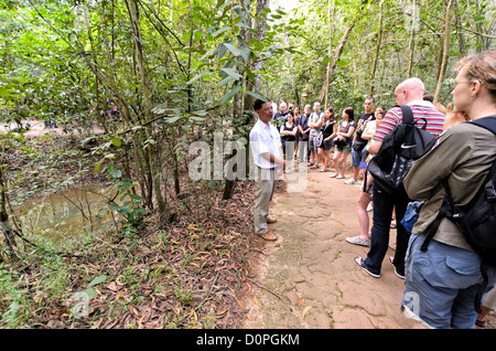 La città di HO CHI MINH, Vietnam - Un tour guida spiega ai turisti che cosa il bombardamento americano ha fatto campagna per il paesaggio circostante. A sinistra del telaio è uno dei tanti bomba crateri che pockmark l'area. I Tunnel di Cu Chi, a nord-ovest di Ho Chi Minh City, erano parte di un più grande galleria sotterranea di rete utilizzato dal Viet Cong nella guerra del Vietnam. Parte dell'originale sistema di tunnel è stata conservata come un attrazione turistica dove i visitatori possono andare verso il basso nello stretto tunnel e vedere presenta sulle precauzioni per la difesa e la vita quotidiana dei vietnamiti che visse e combattute. Foto Stock