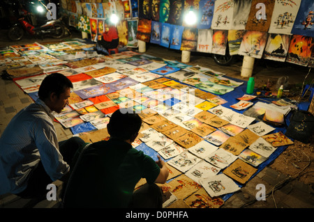 VIENTIANE, Laos - un venditore mostra un cliente che la sua serie di dipinti, disegni e altri illustrato di arte presso il mercato notturno sul Quai Fa Ngum sulle rive del Mekong in Vientiane, Laos. Foto Stock