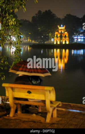 HANOI, Vietnam - una coppia siede sotto un ombrello su una panchina del parco sulle rive del lago Hoan Kiem di notte, con la Torre delle tartarughe (Thap Rua) sullo sfondo. Foto Stock