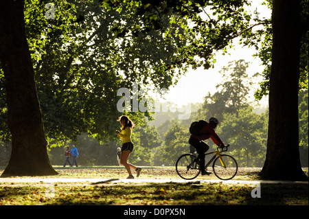 Una giovane donna che fa jogging con le sue sagome, supera un ciclista maschile su un sentiero soleggiato a Hyde Park, Londra, e gli alberi si illuminano in controluce Foto Stock