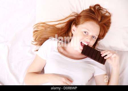 Una giovane donna redhead mangiare il cioccolato a letto. Foto Stock