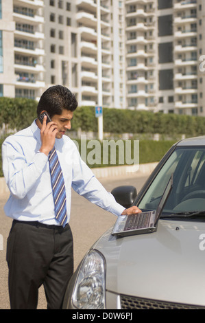 Imprenditore a parlare su un telefono cellulare mentre si utilizza un laptop sul cofano auto, Gurgaon, Haryana, India Foto Stock
