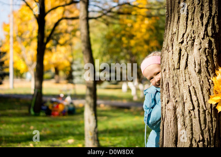 Ragazza giovane giocando un peek boo behimd nascondere il tronco di albero in un parco di autunno e di peering con un commento irriverente fuori dal lato Foto Stock