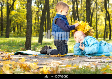 Little Boy facendo una corona di colorati in giallo le foglie di autunno a posto sulla testa del suo ridere sorella giacente a terra accanto a lui. Foto Stock