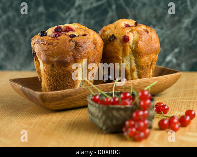 Close-up di due muffin ai mirtilli, su di un piatto di legno con un cestino di ribes rosso nella parte anteriore, sulla sommità di una cucina in legno board Foto Stock