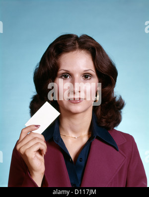 Anni Settanta RITRATTO DI BRUNETTE donna che indossa tuta HOLDING BLANK BUSINESS CARD guardando la fotocamera Foto Stock