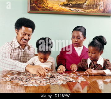 Anni Settanta afro-americano della famiglia uomo donna madre padre di due bambini ragazze seduta tabella facendo Jigsaw Puzzle Retrò Foto Stock