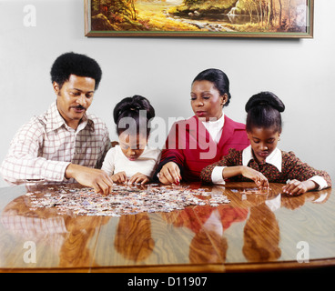 Anni Settanta famiglia americana africana padre MADRE DI DUE FIGLIE seduta a tavola facendo PUZZLE Foto Stock