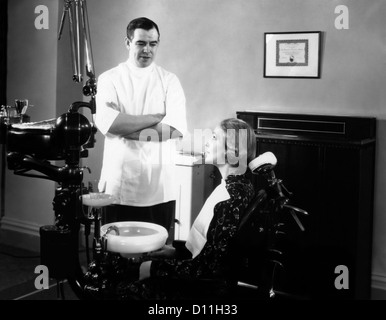 1930s 1940s uomo dentista con le braccia incrociate a parlare con una donna paziente seduto in poltrona odontoiatrica Foto Stock