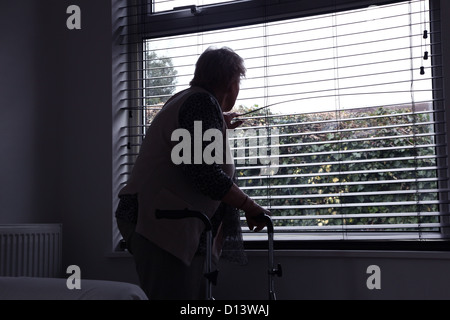 Senior donna con un telaio a piedi guardando fuori da una finestra cieca. Vista posteriore, silhouette. Foto Stock