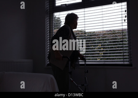 Senior donna sola con un telaio a piedi guardando fuori da una finestra cieca. Posteriore o laterale, silhouette. Foto Stock
