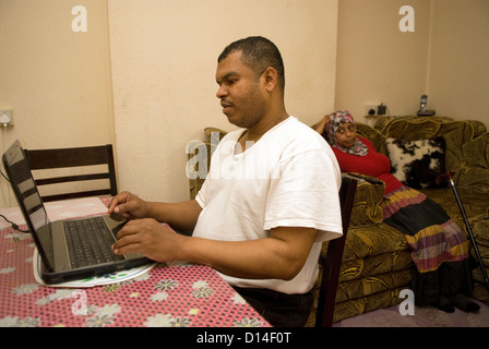 Il marito che si prende cura di sua moglie disabili a casa (seduto, amputazione della gamba) utilizza il computer portatile mentre mia moglie guarda la televisione Foto Stock