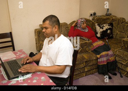 Il marito che si prende cura di sua moglie disabili a casa (seduto, amputazione della gamba) utilizza il computer portatile mentre mia moglie guarda la televisione Foto Stock