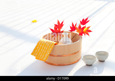 Asciugamano, bottiglia di sake e rosse foglie di acero in un cestello di legno Foto Stock