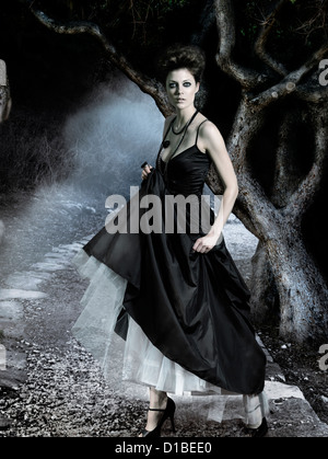 Giovane e bella donna che indossa lungo classico abito nero in una oscura foresta misteriosa. In modo digitale immagine composta. Foto Stock