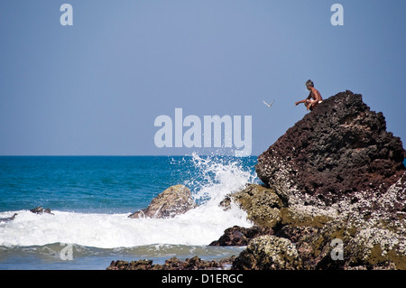 Vista orizzontale di un uomo indiano appollaiato precariamente su una roccia a pesca di Papanasam spiaggia di Varkala, Kerala. Foto Stock