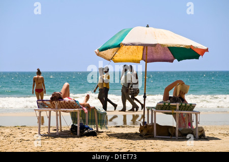 Vista orizzontale della femmina occidentale i turisti a prendere il sole sulla spiaggia di Varkala con un gruppo di Indiani gli uomini passati a piedi. Foto Stock