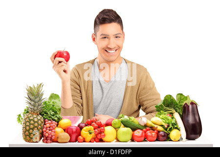 Bel ragazzo sorridente in possesso di un Apple e in posa su un tavolo pieno di verdura e frutta isolato su bianco Foto Stock