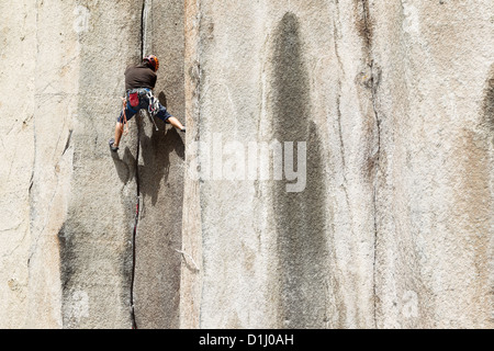 Un arrampicatore che indossa un casco, un'imbracatura e l'attrezzatura da arrampicata sale su un'alta parete di granito verticale. L'arrampicatore è fissato da una fune e si muove attivamente verso l'alto, utilizzando maniglie e punti di appoggio Foto Stock