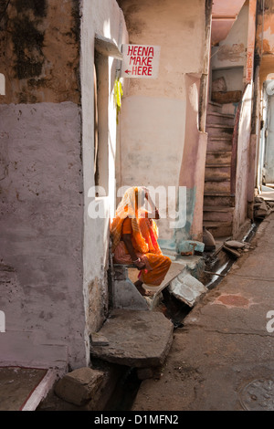 Una donna indiana in un sari arancione si siede sui gradini di una casa pubblicità Henna fare fino in una strada stretta con gradini Foto Stock
