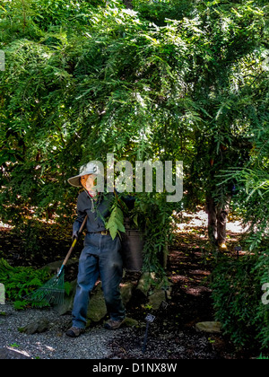 Portando un rastrello e indossando un cappello per il sole, un asiatico giardiniere lavora sotto un albero a New York Giardini Botanici del Bronx. Foto Stock