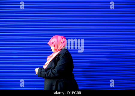 Una donna musulmana guardando il suo telefono cellulare con velo di colore rosa passato a piedi un brillante, blu royal otturatore Foto Stock