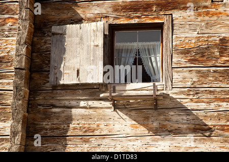 Un vecchio edificio alpino mostra la sua finestra aperta e dentro le sue tende bianche nelle ombre del sole del pomeriggio. Foto Stock
