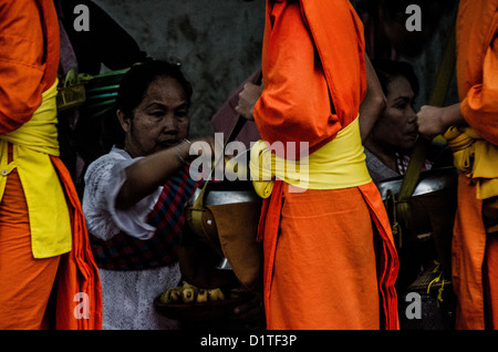 LUANG PRABANG, Laos: Monaci buddisti e novizi in luccicanti accappatoi di zafferano camminano in un unico file per le strade di Luang Prabang durante la cerimonia di consegna delle elemosine del mattino presto nota come tak bat. La gente del posto e i turisti si allineano lungo il percorso, offrendo cibo in questo rituale quotidiano che è centrale per la tradizione buddista del Laos. Foto Stock