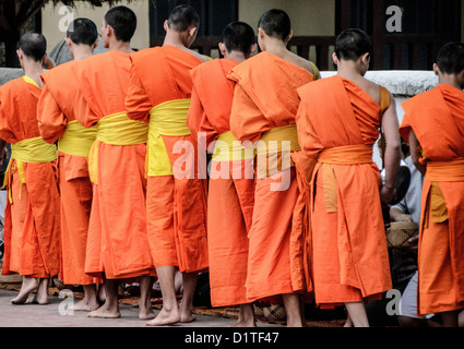 LUANG PRABANG, Laos: Monaci buddisti e novizi in luccicanti accappatoi di zafferano camminano in un unico file per le strade di Luang Prabang durante la cerimonia di consegna delle elemosine del mattino presto nota come tak bat. La gente del posto e i turisti si allineano lungo il percorso, offrendo cibo in questo rituale quotidiano che è centrale per la tradizione buddista del Laos. Foto Stock