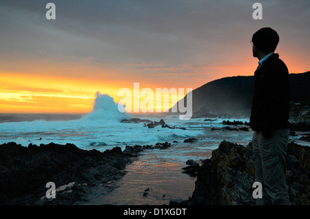 Un uomo guarda il sole di setting e drammatico di onde che si infrangono sulla riva del Sud Africa Foto Stock