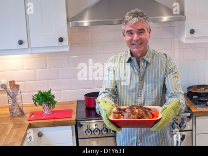 Sorridendo rilassato fiducioso uomo maturo in cucina contemporanea presenta un caldo cucinato al momento pasto di pollo arrosto e verdure Foto Stock