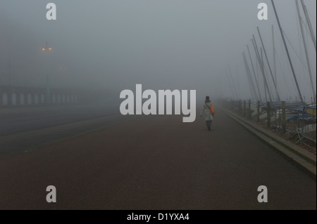 Donna con sacco arancione camminando lungo il lungomare di vuoto nella fitta nebbia Foto Stock