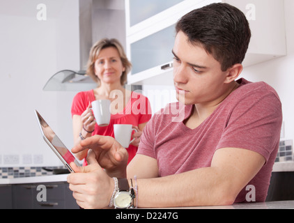 Giovane uomo utilizzando una tavoletta digitale, guardando lo schermo o monitor. Situato in una cucina moderna con una donna che mantiene due tazze sorrisi Foto Stock