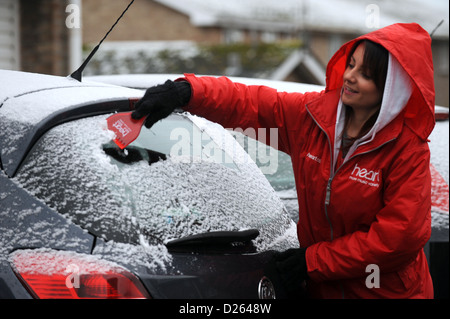 Giovane donna vestita di cuore stazione radio FM giacca la rimozione di neve e ghiaccio da Vauxhall corsa auto utilizzando un raschiatore di cuore Foto Stock