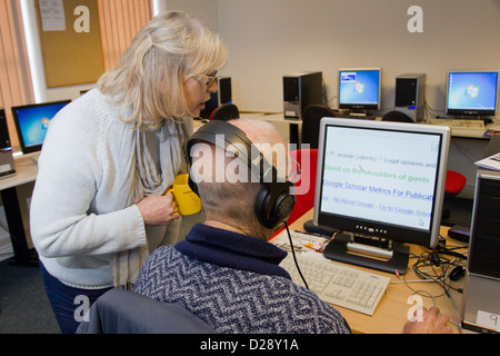Computer di classe per le persone con disabilità visive - volontario con l'uomo utilizzando le cuffie per audio e testo ingrandito. Foto Stock