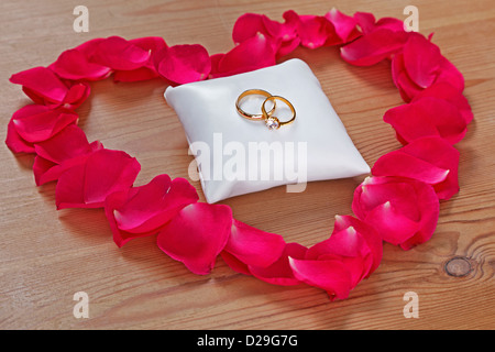 Nozze d'oro e anello di fidanzamento su un piccolo cuscino bianco circondato da un petalo di rosa cuore su uno sfondo di legno. Foto Stock