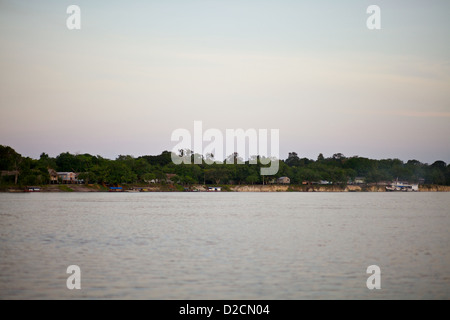 Vista panoramica di un piccolo villaggio sul fiume Rio delle Amazzoni al crepuscolo, circondato da vegetazione lussureggiante e acque calme sotto un cielo pastello in Brasile Foto Stock