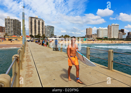 Durban, Sud Africa. Un giovane surfer passeggiate lungo il molo Durbans North Beach. Alberghi e appartamenti sono in background. Durban, Sud Africa. Foto Stock