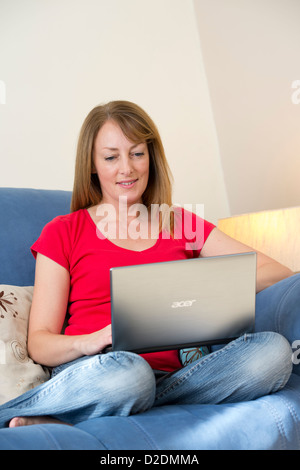 Donna di mezza età utilizzando un computer portatile per navigare in internet mentre seduto nel suo soggiorno a casa. Foto Stock