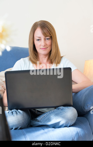 Donna di mezza età utilizzando un computer portatile per navigare in internet mentre seduto nel suo soggiorno a casa. Foto Stock