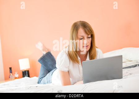 Donna di mezza età utilizzando un computer portatile per navigare in internet mentre giacente sul suo letto. Foto Stock