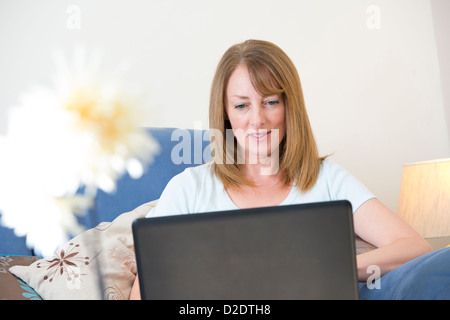 Donna di mezza età utilizzando un computer portatile per navigare in internet mentre seduto nel suo soggiorno a casa. Foto Stock