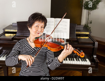 Senior donna adulta a suonare il violino a casa con il pianoforte in background Foto Stock
