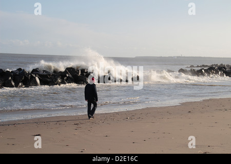 Un uomo che cammina lungo una spiaggia con onde che si infrangono sulle rocce dietro. (Wexford, Irlanda). Foto Stock
