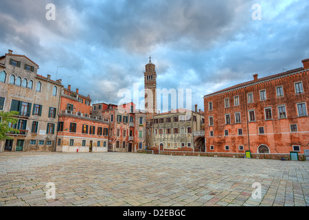 Piccola plaza tra vecchi tipici edifici colorati e alto campanile sullo sfondo sotto il bellissimo cielo molto nuvoloso in serata a Venezia. Foto Stock